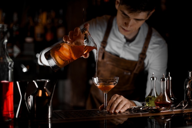 Atrakcyjny męski barman nalewający ciemnego brązowego koktajlu alkoholowego z miarki do szklanki