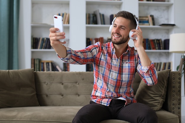 Atrakcyjny mężczyzna biorąc selfie zdjęcie lub własny film z telefonu komórkowego w domu, siedząc na kanapie.