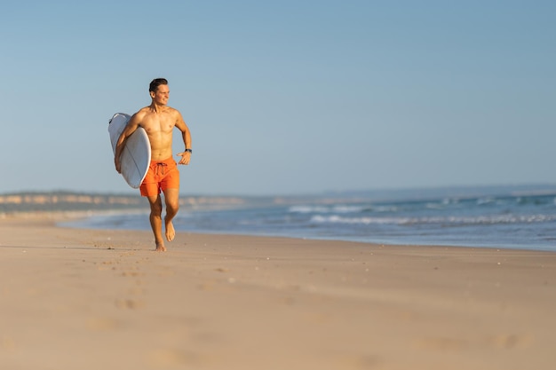 Atrakcyjny mężczyzna o ładnym atletycznym ciele biegający po brzegu z deską surfingową