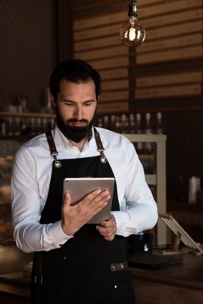 Atrakcyjny mężczyzna w fartuchu barista lub właściciel stoi w kawiarni trzymając tablet