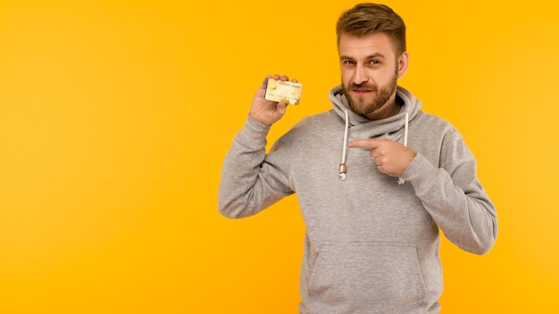 Atrakcyjny mężczyzna w szarej bluzie z kapturem wskazuje palcem na kartę kredytową, która trzyma w ręku na żółtym tle - obraz