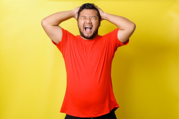Atrakcyjny młody azjatycki grubas w czerwonej koszulce z błyszczącymi zębami krzyczy, dotykając głowy zamykając oko