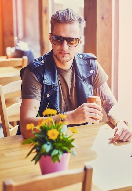 Atrakcyjny młody blond brodaty mężczyzna ubrany w skórzaną kurtkę siedzi przy stole i pije sok.