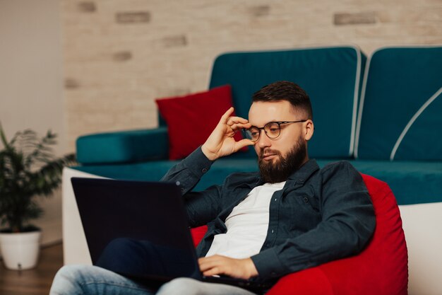 Atrakcyjny młody brodaty poważny mężczyzna w okularach pracuje na fotelu w domu, za pomocą laptopa.