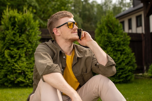Atrakcyjny młody człowiek w ubranie, odwracając wzrok i rozmawiając przez telefon komórkowy, siedząc sam na trawie, wakacje, weekend, taryfy dla operatora połączeń komórkowych, koncepcja komórkowa