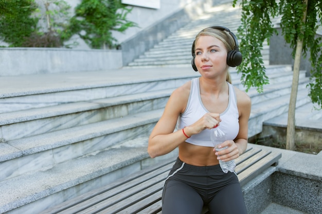 Atrakcyjny młody fitness blond kobieta słucha muzyki na słuchawkach i trzyma butelkę wody na zewnątrz. Koncepcja zdrowego stylu życia