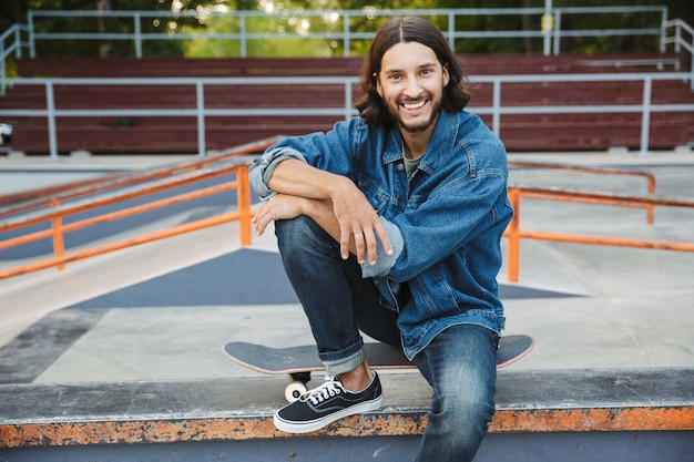 Atrakcyjny młody hipster mężczyzna siedzący w skate parku z deskorolką