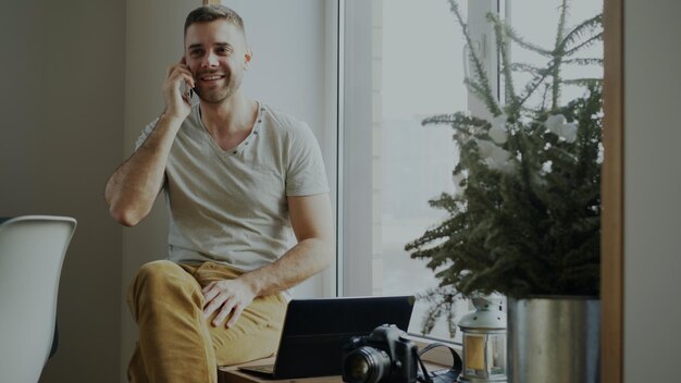 Atrakcyjny młody mężczyzna rozmawiający telefon siedzący na parapecie z laptopem i aparatem w domu