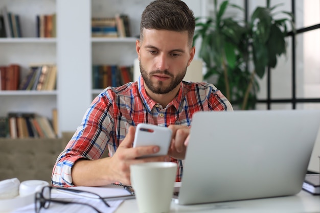 Atrakcyjny młody mężczyzna siedzi przy biurku w domowym biurze i za pomocą telefonu komórkowego do sprawdzania sieci społecznościowych.