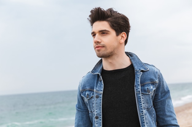 Atrakcyjny młody mężczyzna ubrany w dżinsową kurtkę spacerujący po plaży