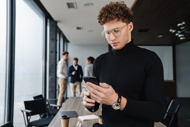 Atrakcyjny młody pewny siebie biznesmen w stroju wizytowym siedzi w biurze ze swoimi kolegami na ścianie, używając telefonu komórkowego