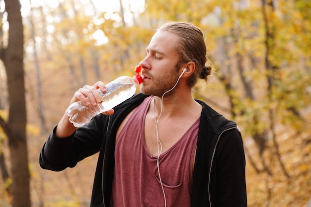 Atrakcyjny młody sportowiec biegający w jesiennym lesie, słuchający muzyki przez słuchawki, trzymający butelkę wody