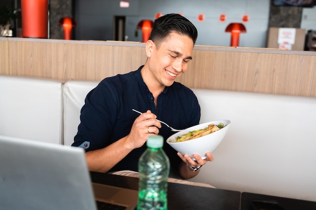 Atrakcyjny młody szczęśliwy mężczyzna siedzący i jedzący sałatkę Komputer laptop i butelka wody na stole