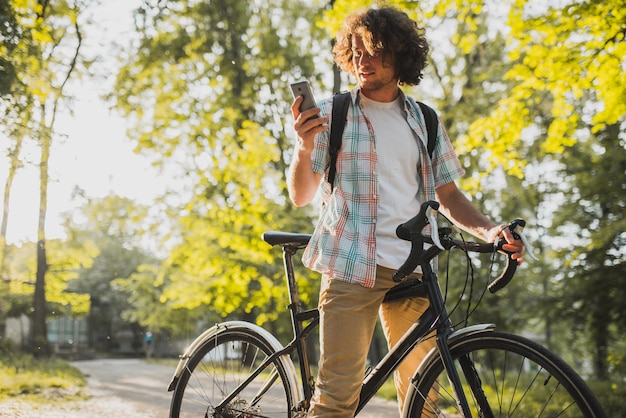 Atrakcyjny młody szczęśliwy mężczyzna z kręconymi włosami patrząc na swój telefon komórkowy wybierając aplikację gps na drogę na rower Szczęśliwy uśmiechnięty kolarz z plecakiem surfowanie po internecie za pomocą smartfona