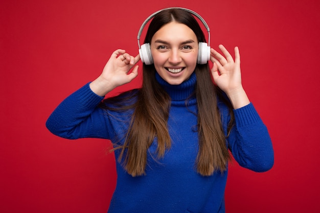 Atrakcyjny Pozytywny Uśmiechający Się Młoda Brunetka Kobieta Ubrana W Niebieski Sweter Na Białym Tle Nad Czerwonym Tle ściany Noszenie Białych Słuchawek Bluetooth, Słuchając Fajnej Muzyki I Bawiąc Się Patrząc Na Kamery