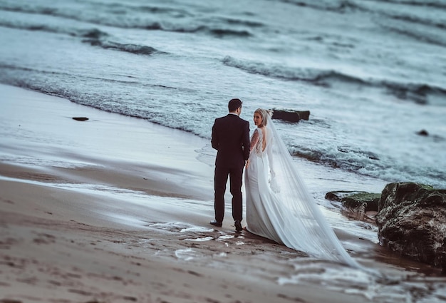 Atrakcyjny ślub Para Trzyma Się Za Ręce Spacerując Po Plaży W Pobliżu Fal Oceanu W Wieczornym świetle.