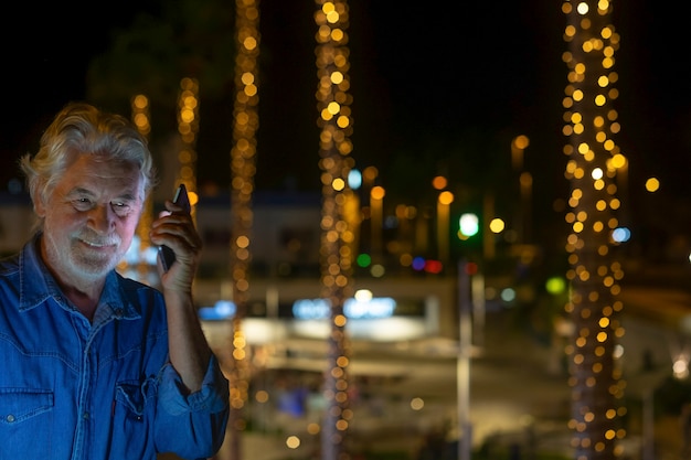 Atrakcyjny starszy mężczyzna korzystający z telefonu komórkowego na zewnątrz w nocy w pobliżu oświetlonej choinki