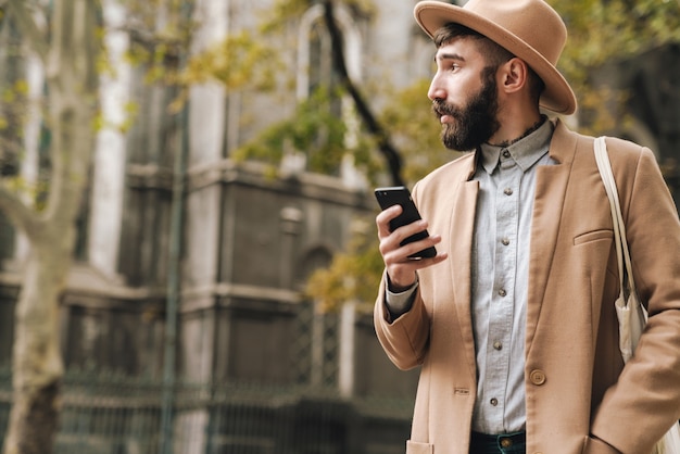 atrakcyjny stylowy młody mężczyzna w kapeluszu trzymającym telefon komórkowy podczas chodzenia na zewnątrz