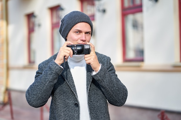 Zdjęcie atrakcyjny, uśmiechnięty hipster modny mężczyzna ubrany w szary płaszcz, biały sweter i szary kapelusz z aparatem