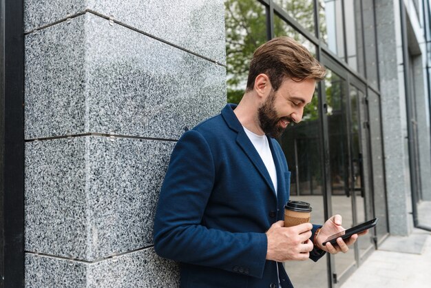 Atrakcyjny Uśmiechnięty Młody Brodaty Mężczyzna Ubrany W Kurtkę, Używający Telefonu Komórkowego, Stojący Na Zewnątrz W Mieście I Pijący Kawę Na Wynos