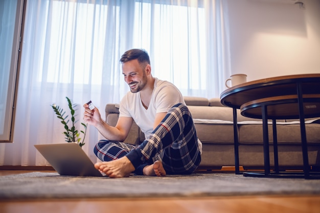 Atrakcyjny Uśmiechnięty Nieogolony Kaukaski Mężczyzna W Piżamie Siedzi Na Podłodze Boso, Trzyma Kartę Kredytową I Korzysta Z Laptopa Do Płatności Online.