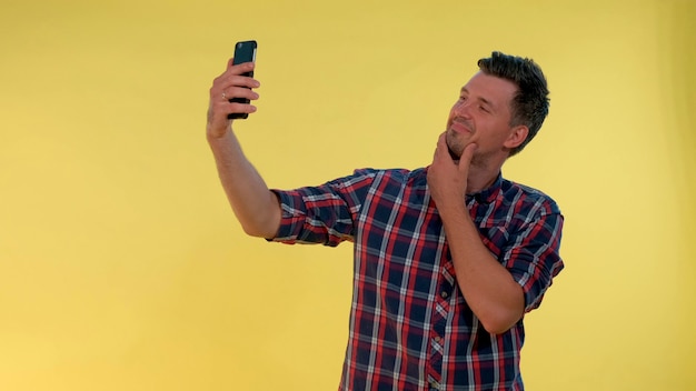 Atrakcyjny wysoki chłopak robi selfie na smartfonie, uśmiechając się i stojąc na żółtym tle