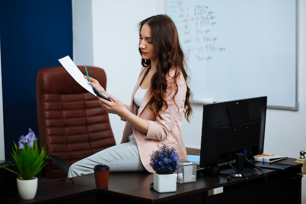 Atrakcyjny życzliwy bizneswoman przy pracą w biurowym obsiadaniu przy biurkiem z papierkową robotą i komputerem stacjonarnym ono uśmiecha się przy kamerą