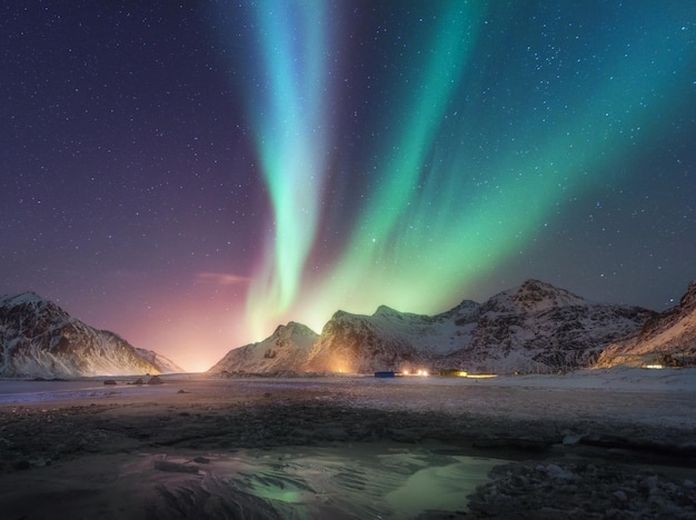 Aurora borealis nad zaśnieżoną górą i piaszczystą plażą zimą w nocy Zorza polarna na Lofotach Norwegia Niebo z gwiazdami i zorzą polarną Krajobraz ze światłami miasta wybrzeża morskiego zorzy polarnej