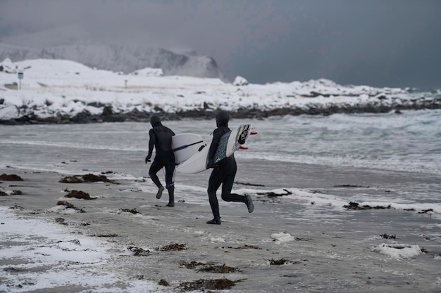 Autentyczni lokalni arktyczni surferzy biegający na zaśnieżonej plaży po surfowaniu w Morzu Norweskim. Ośnieżone górskie tło w zimie. Norweskie Lofoty. Wybrzeże Morza Norweskiego. Aktywna woda zimowa