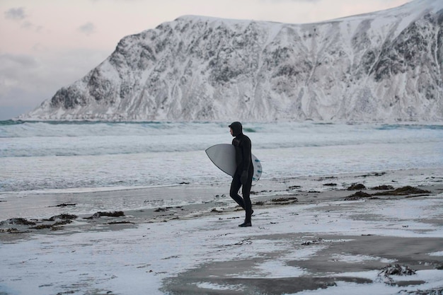 Autentyczny lokalny surfer arktyczny idący po plaży po surfowaniu na Morzu Północnym. Wybrzeże Morza Norweskiego. Sporty ekstremalne w wodzie zimą