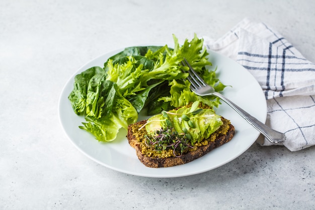 Avocado toast z pesto, kiełkami i sałatką na białym talerzu.