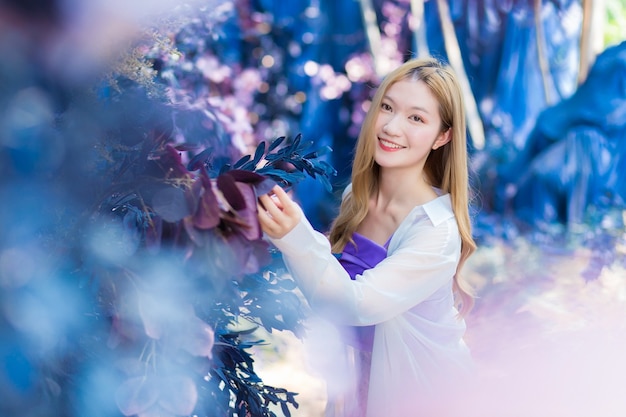 Azjatka, która ma brązowowłose włosy, nosi białą koszulę i fioletową koszulę, uśmiecha się w niebieskim kwiatowym ogrodzie