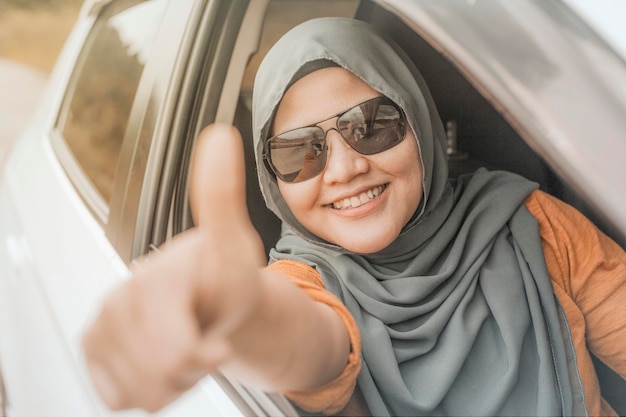 Azjatka muzułmańska uśmiecha się podczas jazdy, bawi się w wycieczce wakacyjnej, jeździ samochodem, dzieli kciuki w górę.