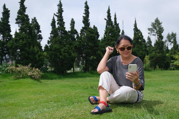 Zdjęcie azjatka siedząca na trawie w parku i dzwoniąca do telefonu.