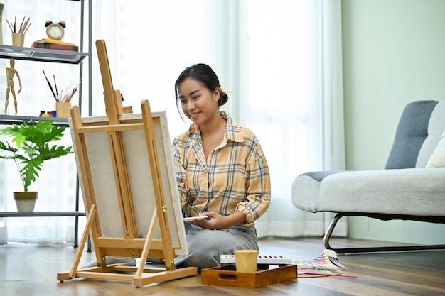 Azjatka siedzi na podłodze w salonie malując farbą akrylową na sztalugach płóciennych