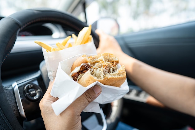 Azjatka trzymająca hamburgera i frytki do jedzenia w samochodzie jest niebezpieczna i ryzykuje wypadek