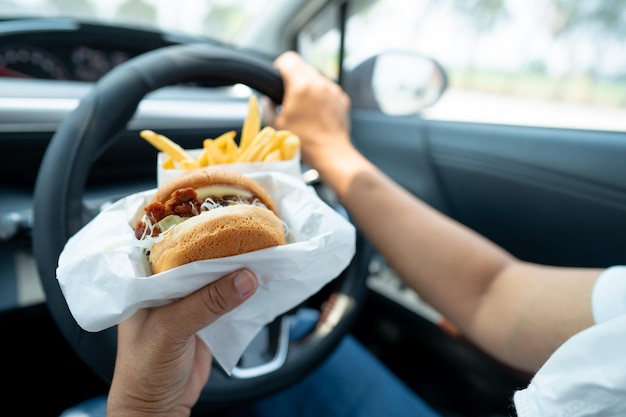 Azjatka trzymająca hamburgera i frytki do jedzenia w samochodzie jest niebezpieczna i ryzykuje wypadek