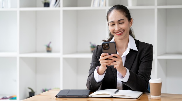 Azjatka w formalnym garniturze siedząca w biurze wesoła podczas korzystania ze smartfona i pracy