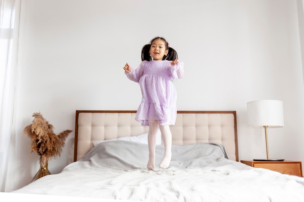 Azjatka w sukience cieszy się i skacze na łóżku w domu Koreańskie dziecko w uroczystym stroju tańczy
