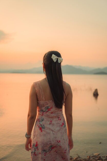 Azjatka w sukience z spokojnym nastrojem na brzegu jeziora w zachodzie słońca