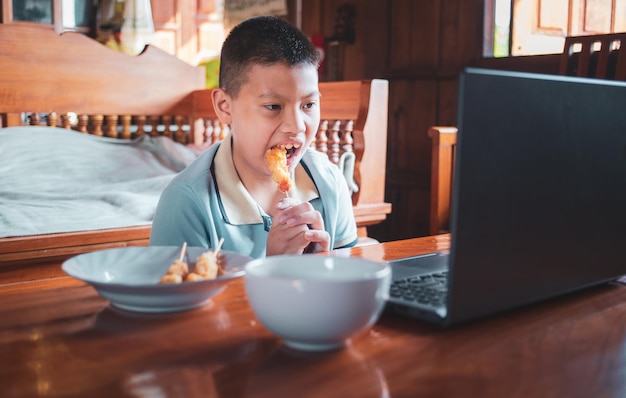 Azjatyccy chłopcy jedzą śmieciowe jedzenie, siedząc przy biurku z notebookami i laptopami w domu
