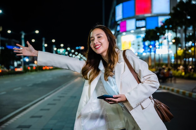 Azjatycka biznesmenka idąca na grad machająca ręczną taksówką na drodze na ulicy miasta w nocy, piękna kobieta uśmiechająca się za pomocą aplikacji na smartfona, witająca się z ręką w górę, dzwoniąca do taksówki na zewnątrz po późnej pracy