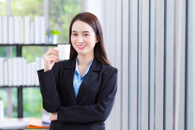 Azjatycka biznesowa dama, ubrana w niebieską koszulę i czarny garnitur, trzyma w dłoni filiżankę kawy i uśmiecha się