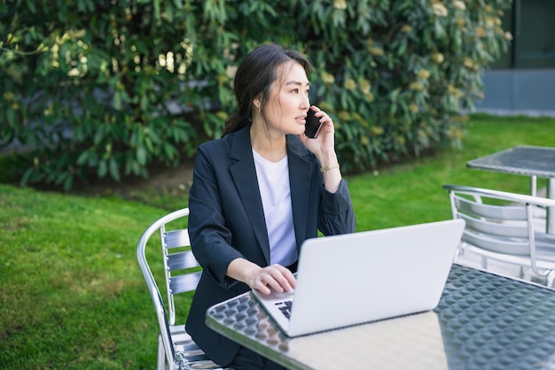 Azjatycka biznesowa kobieta opowiada na telefonie i pracuje z laptopem plenerowym