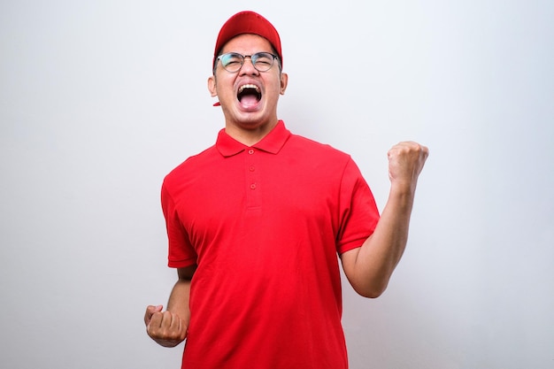 Azjatycka dostawa mężczyzna ubrany w czapkę stojącą nad odizolowanym czerwonym tle, bardzo szczęśliwy i podekscytowany, wykonując gest zwycięzcy z podniesionymi rękami, uśmiechając się i krzycząc o sukces Koncepcja uroczystości