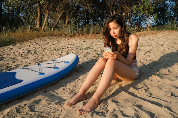 Azjatycka dziewczyna siedzi na plaży na piasku z telefonem i surfuje po internecie