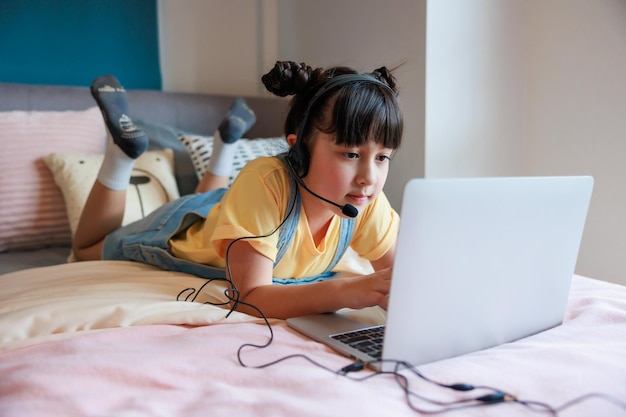 Azjatycka dziewczynka korzystająca ze słuchawek i laptopa uczy się online z nauczycielem wideo w sypialni do nauki na odległość