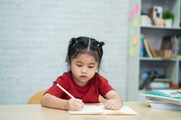 Azjatycka dziewczynka ubrana w czerwoną koszulę pisze notatki w zeszycie i czyta książkę do nauki online na drewnianym biurku w salonie w domu Koncepcja edukacji online z domu