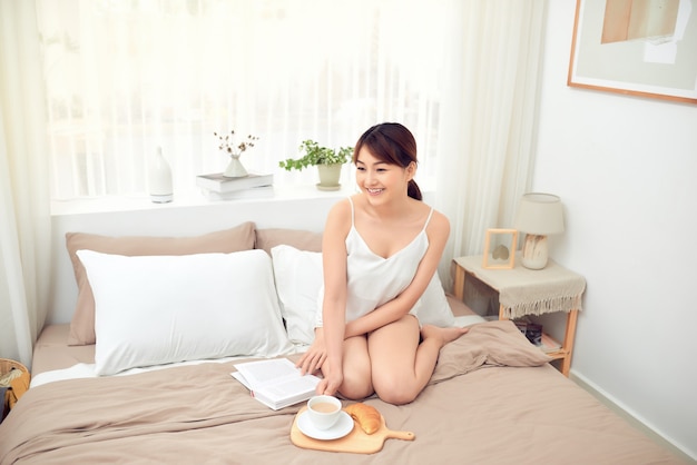 Azjatycka kobieta czytająca książkę z filiżanką kawy w łóżku rano