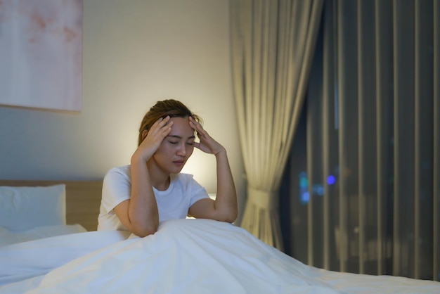 Azjatycka kobieta leżąca na białym łóżku w sypialni wyglądająca na zmartwioną lub myślącą o swoim życiu lub pracy w środku nocy w domu